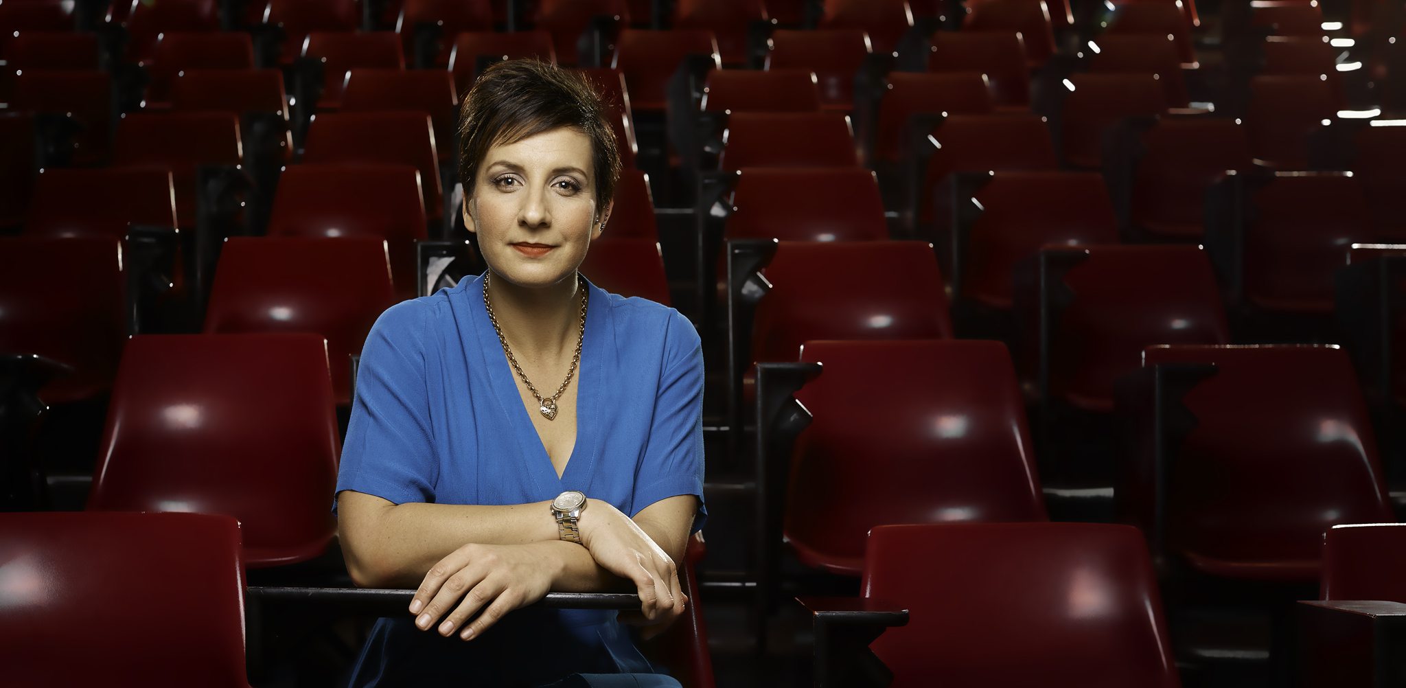 Naomi Malouf seated in an empty university lecture theatre, rows of red seats behind her